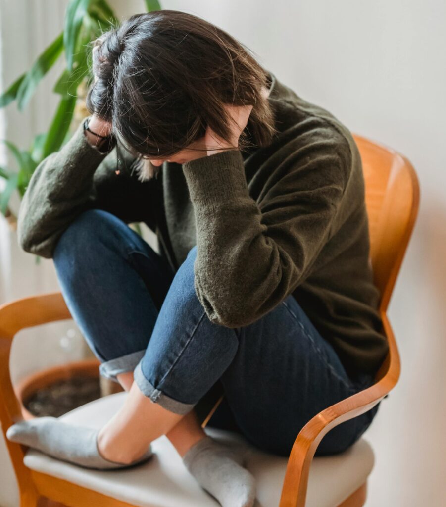 Full body of unrecognizable young worried female in casual clothes sitting on chair with crossed legs and covering ears with hands at home